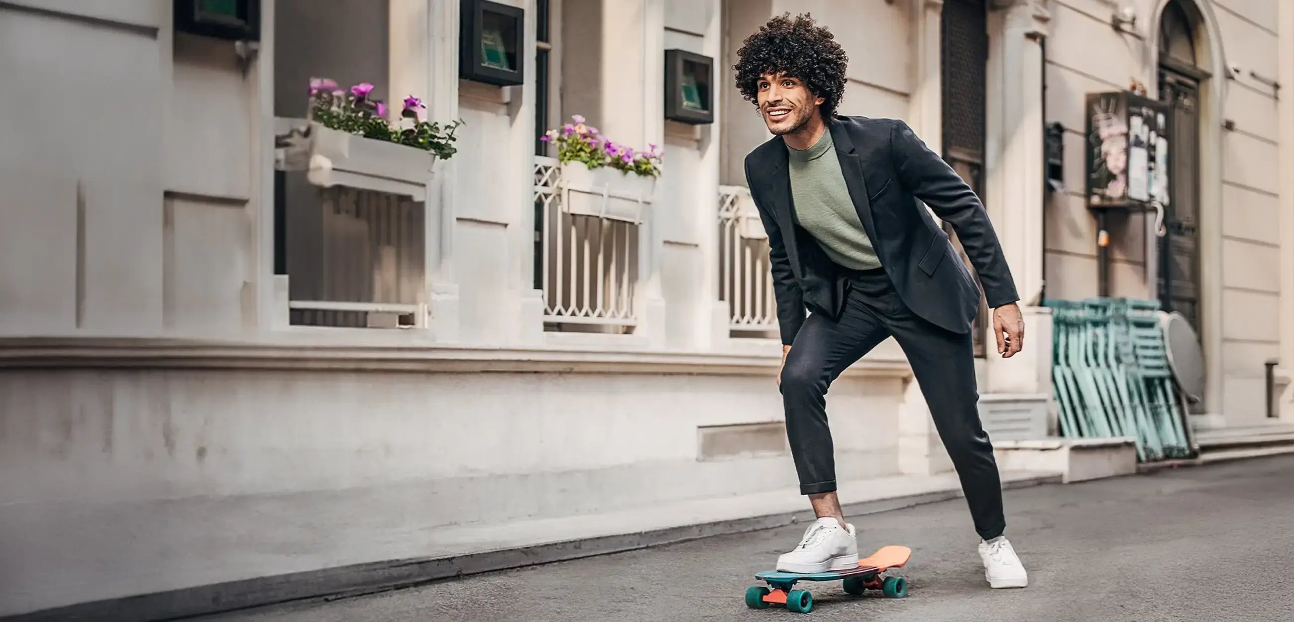 Ein Mann in einem Mietanzug von adretto fährt auf einem Skateboard eine Strasse entlang.